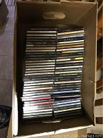 Top view of a cardboard box filled with approximately 50 music CDs in jewel cases showing various artist names on spines