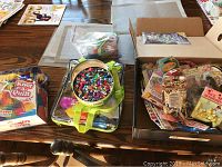 Craft supplies on a table including a metal tin filled with colorful beads, a clear bag with beads, and a cardboard box with multiple craft kits and supplies in original packaging.