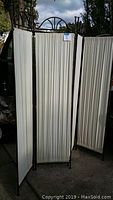 Three black wrought iron framed folding panels with off-white cloth panels arranged outdoors, showing full view of panels and overall condition