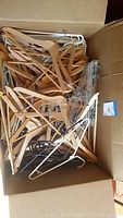 Box filled with many wooden and plastic hangers of different styles and colors, mostly natural wood finish and white plastic.