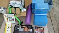Box with blue foam yoga blocks, purple yoga mat, several exercise journals and fitness DVDs, and green yoga straps.