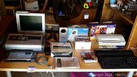 Wide view of various office electronics and multimedia devices including two printers, virtual reality glasses, cordless keyboards, and multiple DVD players and accessories laid out on a table.