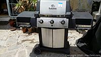 Front view of Broil King Signet barbecue grill with stainless steel doors and control panel, featuring 3 control knobs and black side shelves on a stone patio.