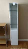 Tall white Ikea bathroom shelf with glass shelves seen from the front.