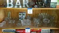 Shelf showing multiple clear glass décor pieces including candlestick holders, candy dishes, napkin holders, and vases in various sizes and shapes