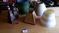 Two lampshades and two table lamps visible on wooden surface, one green cylindrical lampshade and three fabric lampshades in beige and white striped pattern