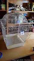 Front and side view of metal birdcage with scrollwork and plastic base on a wooden table with miscellaneous items in the background.