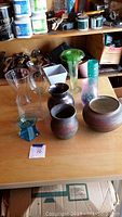 Seven flower vases arranged on a wooden table showcasing both glass and pottery materials. Pots have earthy brown tones, and green and clear glass vases are present. Miscellaneous workshop items are visible in the background.