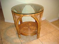 Front angle view of round wicker patio table with thick glass top and leaf patterned shelf