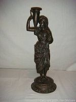 Full view of antique metal candlestick showing Middle Eastern lady holding vessel above shoulder, standing on decorated base.