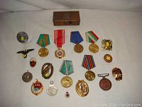 Photo of the entire lot showing 17 military medals, badges, and a rusted metal box, arranged on white fabric.