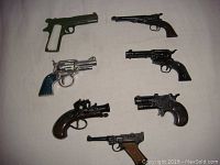 Photo showing 7 vintage metal cap guns laid out on a white cloth, including pistols and revolvers of various styles and colors.
