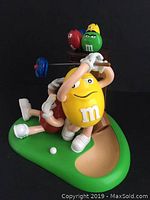 Front view showing yellow M&M character swinging golf club with green and red M&M characters near golf bag on green base