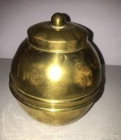 Top and side view showing the spherical brass caddy with removable lid and embossed text and lion figure.