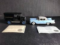 Side view of the two diecast cars - 1924 Chevy Hi-Ton Police Patrol truck in black with blue wheels on left, 1958 Cadillac Eldorado light blue and white on right, both with certificates of authenticity in front.