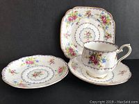 Photo showing the teacup on its saucer, an additional saucer, and one square side plate, all with floral and gold designs.