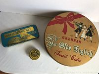 Three vintage tins shown on a white surface: Grennan Ye Olde English Fruit Cake round tin with decorative bow and people image, small Bo Gin Pills tin, and blue-green rectangular Buffalo Tic Set tin.
