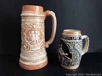 Photo of two German beer steins, an 11 inch tall orange tan stein and a 7 inch tall black and cream colored stein with a couple and German text.