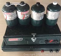 Four propane cylinders positioned on top of a Coleman camp stove with two control knobs and a latch on front.