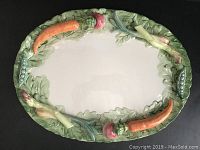 Top view of white oval ceramic platter with raised colorful vegetables (carrots, peas, radishes, green leaves) around rim.