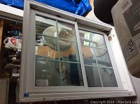 Double pane sliding window with white frame and grid pattern, stored indoors with a fan visible behind it.