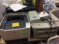 HP LaserJet P2015dn on left and Brother DCP-7020 on right together on wooden surface, with handwritten sticky notes and cables visible