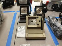 Photo showing three metal passport maker machines used for making or punching passport photos or documents, two dark metal with red levers and one beige with black knobs.