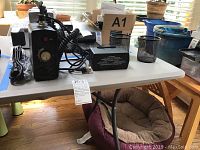 Two black external camera power packs, two black metal mesh pen holders, assorted cables on white folding table indoors near window.