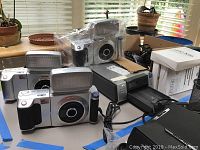 Wide view showing three Sony cameras with large built-in flashes, a Sony digital printer, and other accessories on table.