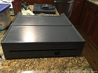 Photo of a black metal POS cash drawer with a key lock on the front, placed on a granite countertop.