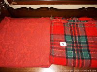 Two folded tablecloths on a wooden surface. Left is a solid red embossed tablecloth, right is a green and red plaid tablecloth with gold accents.