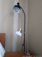 Photo depicting a brown metal floor lamp with a black adjustable neck and white inner lamp shade standing next to a bed, alongside a silver tone desk lamp with a curved neck and round white shade on a desk or table.