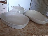 Three white ceramic serving dishes on countertop including French white oval dish, Mikasa covered casserole dish with lid, and large oval serving platter