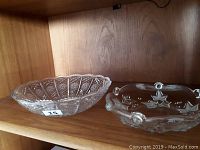 Photo showing two small clear decorative glass serving bowls on a wooden shelf. One bowl has a diamond-cut pattern, the other has stars and small handles.