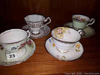 Four porcelain teacups with matching saucers displayed on a wooden shelf, each featuring unique floral patterns and gold accents. One cup and saucer set is mint green, others are white with colorful flowers.