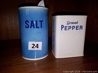 Photo of two vintage salt and pepper shakers by Betty Saint John. Blue cylindrical salt shaker labeled 'SALT' and white square pepper shaker labeled 'Ground PEPPER'.