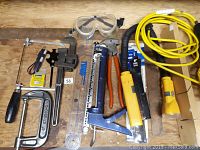 Overview of assorted hand tools on a wooden floor including pipe wrench, clamps, saw, pliers, stapler, ruler, goggles, tape measure, flashlight, and yellow extension cord