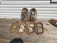 Three pairs of vintage wooden bear claws snowshoes laid out on wooden decking near white siding, showing full frame, leather straps, and footbed made from rawhide or similar material.