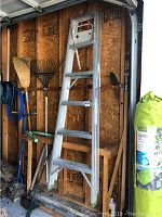 Aluminum ladder standing folded against a wooden wall in a garage, with garden tools around it.