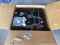 Turntable inside an open cardboard box showing the device with cables and instruction manual on top.