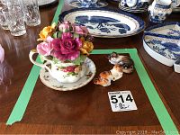 Photo of Royal Albert Old Country Roses teacup music box with floral ceramic top, on wooden table with crystal glasses in background and two ceramic cat figurines to right side.