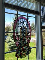 Stained glass peacock panel hanging in window, showing full oval shape and detail with background outdoors