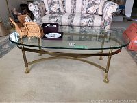 View showing oval glass top coffee table with brass metal legs and decorative claw feet, placed on beige carpet in living room setting.