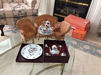 Full view of three wicker miniature chairs arranged around a round wicker table holding a porcelain doll tea set. Nearby are porcelain plates in a box.