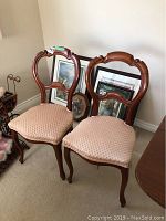 Pair of wooden chairs with upholstered padded seats in light pink patterned fabric. Chairs have carved wood frames with a curved top rail and cabriole legs. Arranged on carpeted floor with framed artwork in the background.