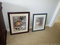 Two framed prints leaning against wall showing winter scenes, one larger with dark wood frame, one smaller with black frame and white mat.