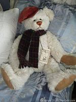 Full image of white teddy bear wearing plaid scarf and red hat on bed showing overall condition and size.