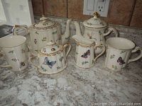 Tea set items including two teapots, one sugar bowl, one creamer, and two mugs, all with butterfly and floral designs and gold trim.