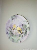Front of a decorative plate showing American goldfinches on a nest among painted leaves in pastel colors