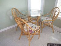 Set of round glass top table with rattan base and four rattan chairs with floral cushions in a room corner.